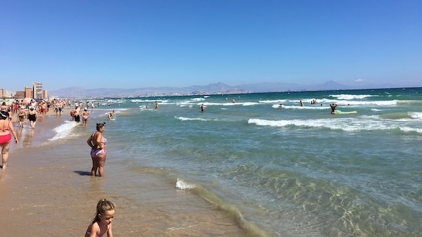 A crowded beach with people walking and surfing, clear skies, and distant mountains.