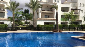 A swimming pool surrounded by a fence, with palm trees and a building with balconies in the background.