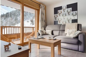 Un salon moderne avec un canapé gris, une table basse en bois et une vue sur un paysage montagneux enneigé, visible à travers de grandes fenêtres.