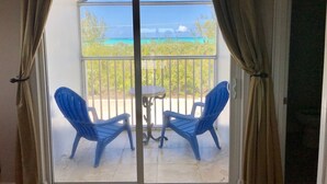 A balcony with two blue chairs and a small table, overlooking a turquoise sea and greenery.