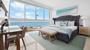 A bedroom with a large window overlooking the sea, a bed with a grey headboard, a desk with a chair, and a bedside table with a lamp. - Heaven can wait,I found a paradise with Ocean view (Noord)