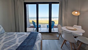 A hotel room with a bed, a dining area with chairs and a table, and a view of the beach through a sliding door. - Beachfront Studio | Ground-Floor Patio + Ocean Views | Steps to Sand | Free Activities Included (Daytona Beach Shores)