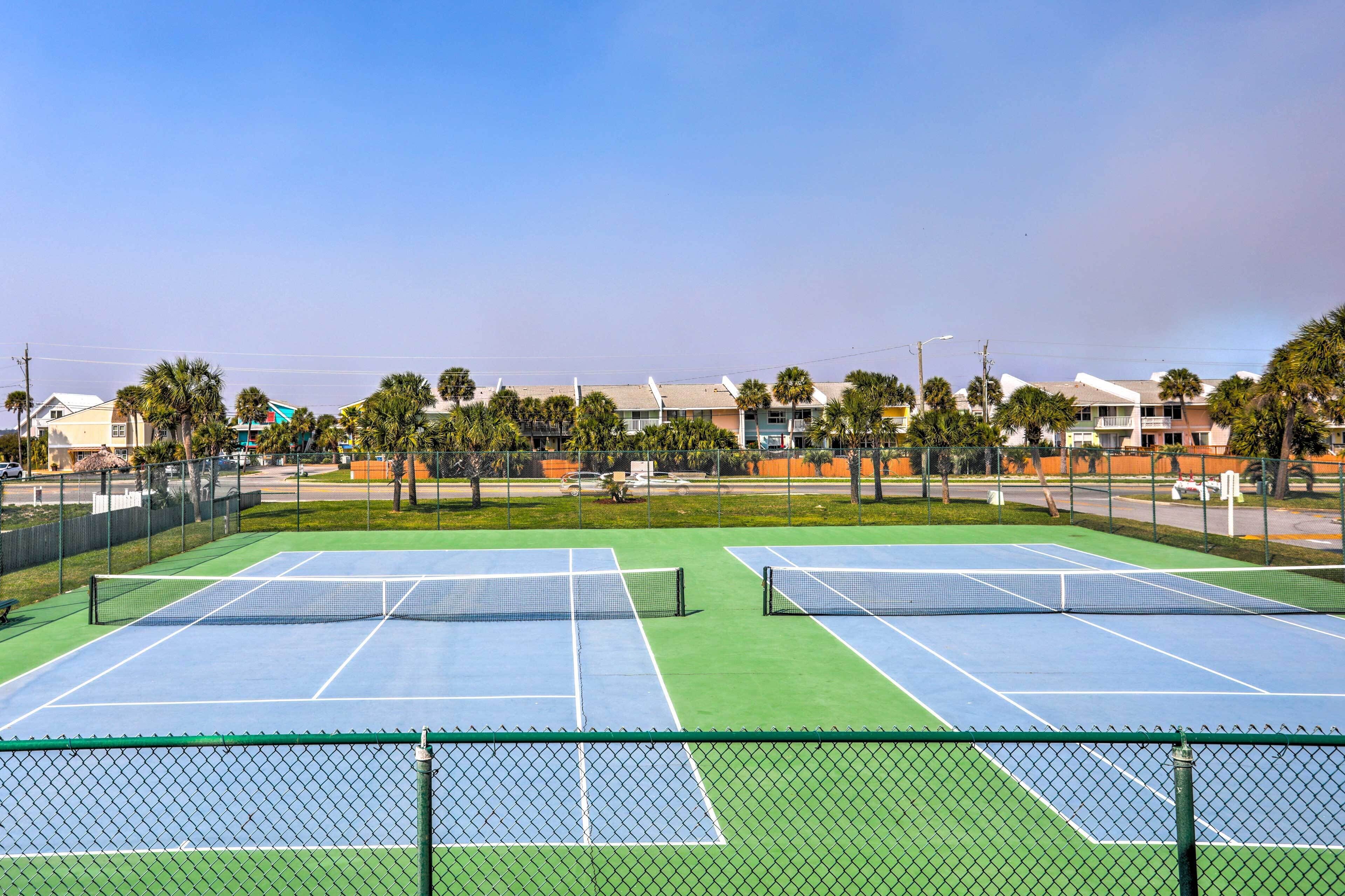 A tennis court with two courts, surrounded by a fence, palm trees, and buildings in the background.