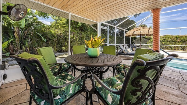 An outdoor patio with a round table and chairs, surrounded by lush greenery and a view of a swimming pool. - Specials - 3BR with Pool, Walk to Town (Cape Coral)