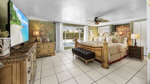 A spacious bedroom with a large bed, a television, a ceiling fan, and a view of the outdoors. - Specials - 3BR with Pool, Walk to Town (Cape Coral)