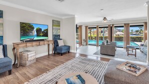 Een ruime woonkamer met een grote flatscreen-tv, comfortabele zitplaatsen, een houten salontafel en uitzicht op een zwembad door grote ramen.