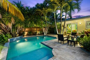 Ein Schwimmbecken, umgeben von üppiger Vegetation und Palmen, mit einer Terrassenfläche, die Outdoor-Möbel und einen gut beleuchteten Innenbereich durch eine Glasdoor bieten.