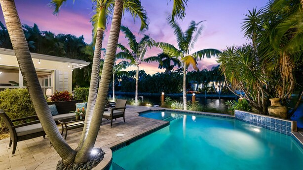 A swimming pool with palm trees, a patio area with seating, and a view of a body of water at dusk. - Charming Florida Home on the Waterway with Coastal Charm and Easy, Relaxed Vacation Vibes! (Wilton Manors)