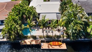 An aerial view of a resort with a swimming pool surrounded by palm trees, a wooden deck with lounge chairs, and a boat docked at the water's edge. - Charming Florida Home on the Waterway with Coastal Charm and Easy, Relaxed Vacation Vibes! (Wilton Manors)