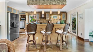 A modern kitchen with a stainless steel refrigerator, a microwave, and a built-in oven. There are three wicker bar stools with cushioned seats. The kitchen features a tiled backsplash and pendant lighting. - Charming Florida Home on the Waterway with Coastal Charm and Easy, Relaxed Vacation Vibes! (Wilton Manors)