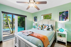 Ein Schlafzimmer mit Bett, Deckenventilator, Nachttisch, Lampe und Fenster mit Blick auf einen Swimmingpool und Palmen.