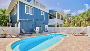 A two-story building with a swimming pool, lounge chairs, and a white fence.