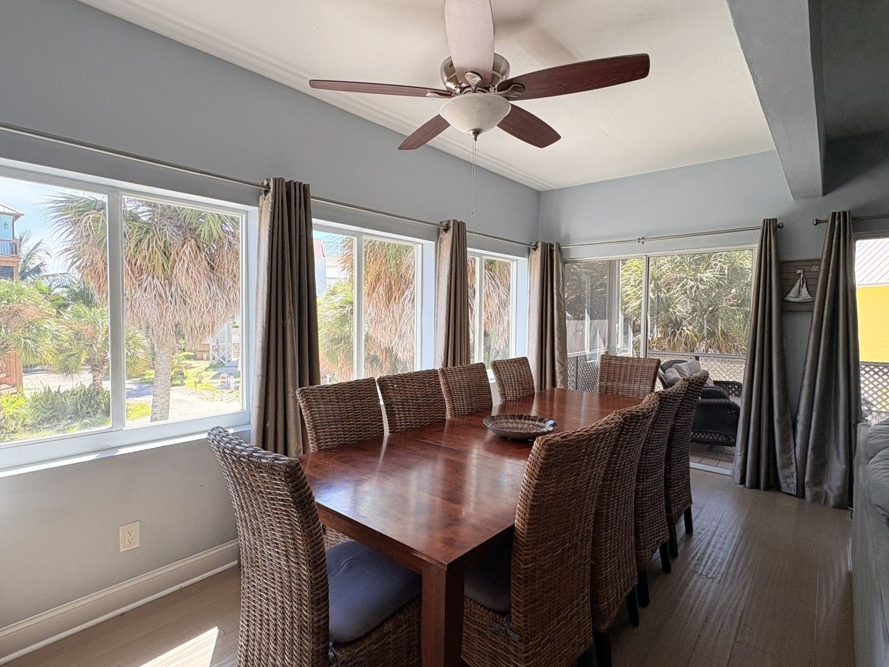 A dining area with a wooden table and wicker chairs, large windows with curtains, and a ceiling fan.