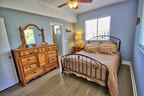 Um quarto com cômoda de madeira, cama com estrutura de metal, ventilador de teto e janela com persianas.