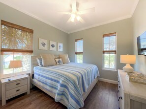 Un dormitorio con cama, mesitas de noche, ventilador de techo y ventanas con persianas.