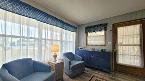 Een kamer met twee blauwe fauteuils, een houten bijzettafeltje met een lamp, een blauwe kast en een groot raam met lichte gordijnen.