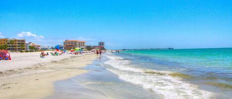 Una playa con agua azul cristalina, un muelle y edificios al fondo.