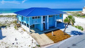Casa de playa de dos pisos con techo azul y paredes blancas, que cuenta con un porche cubierto y un pequeño jardín delantero.