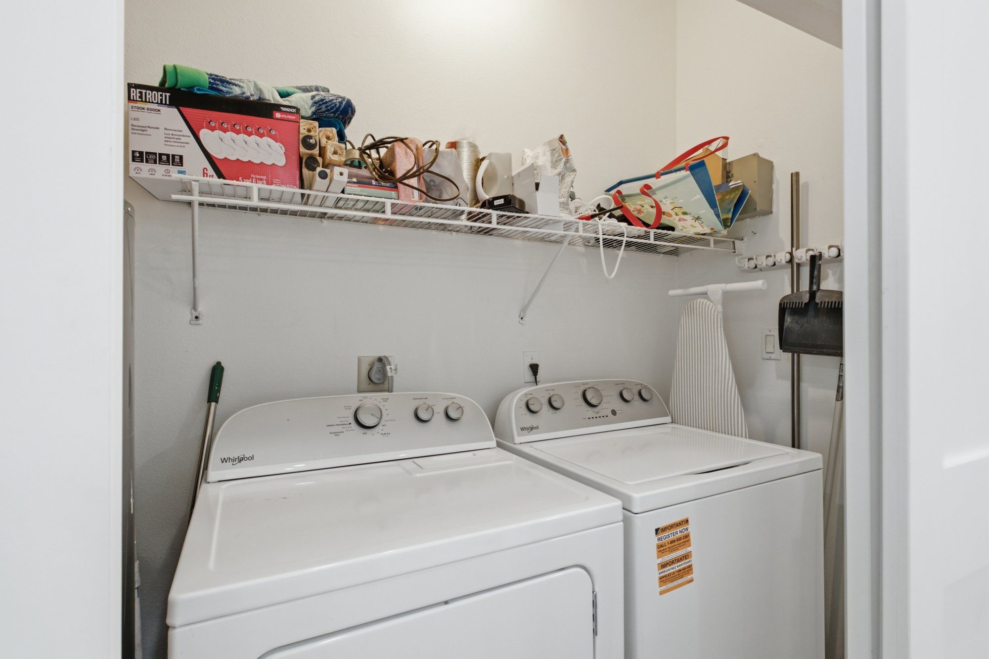 Un área de lavandería con dos lavadoras Whirlpool blancas, un estante superior con varios objetos y un tendedero con ropa.