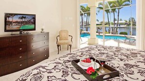 Une chambre d’hôtel avec un grand lit, un téléviseur posé sur une commode en bois, une chaise, une table à manger avec un plateau de nourriture et de boissons, et un balcon donnant sur une piscine et des palmiers.