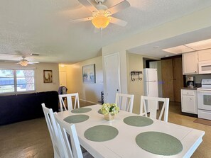 A dining area with a white table, green placemats, white chairs, a ceiling fan, and a kitchen with white appliances.