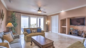 A spacious living room with a ceiling fan, a large window, a flat-screen TV, and a fireplace. - Riverfront Titusville Home w/ Pool + Pickleball! (Titusville)