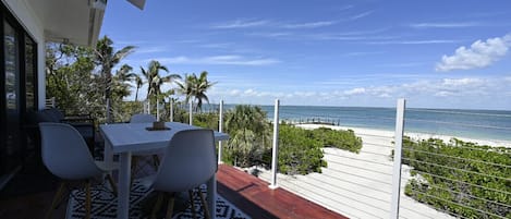 A balcony with a white table and chairs, overlooking a beach with palm trees and a clear sky.