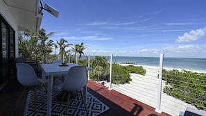 A balcony with a white table and chairs, overlooking a beach with palm trees and a clear sky.