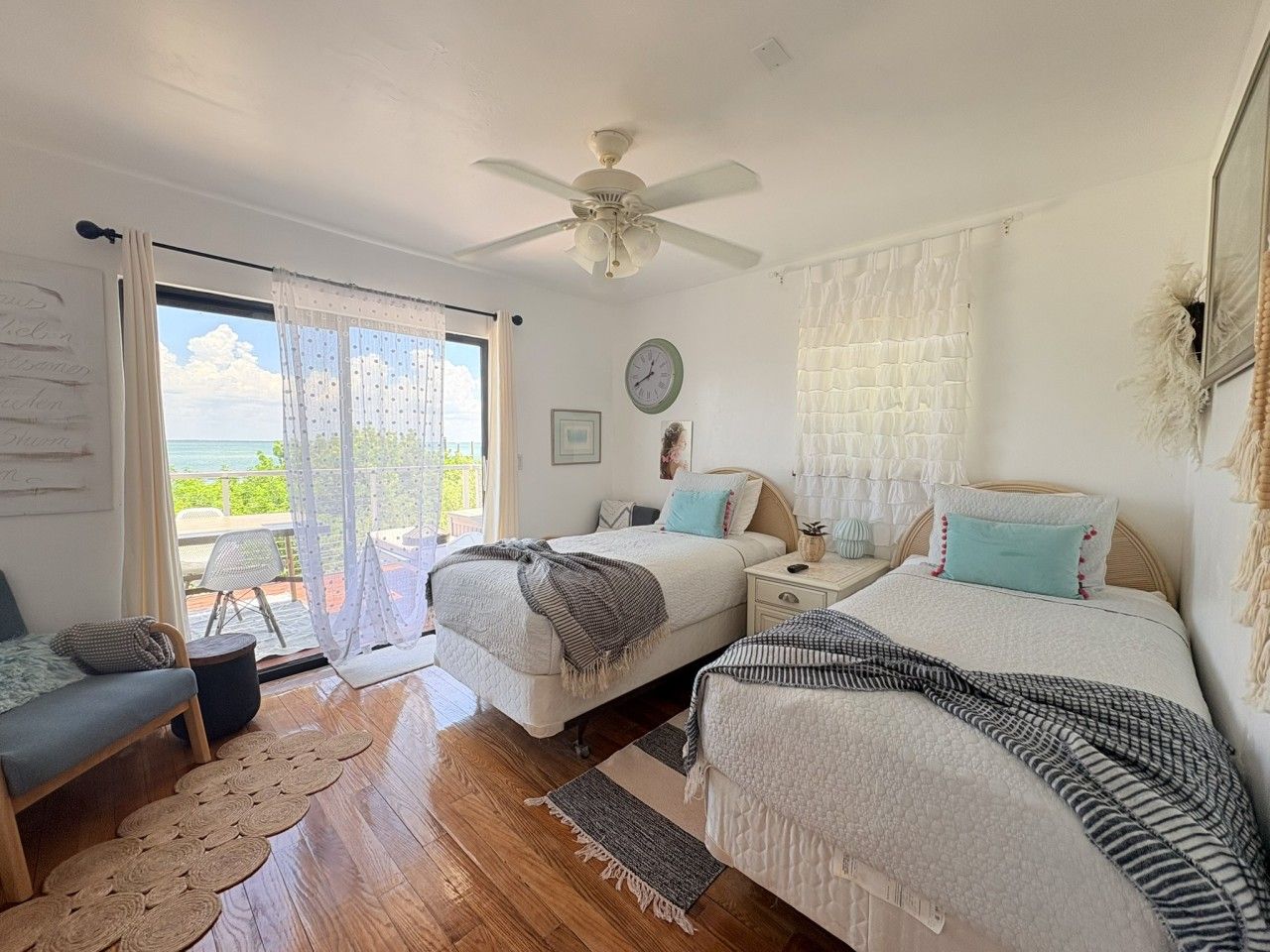 A bedroom with two beds, a ceiling fan, a view of the outdoors, and a large window with curtains.