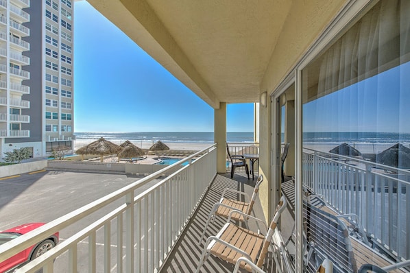 A balcony with a view of the beach and ocean, a pool area, and a red car in the parking lot.