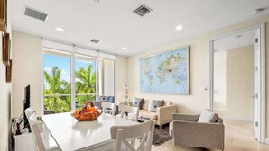 A modern living room with a dining area, large windows, a world map on the wall, and a view of palm trees outside. - Hemingway Condo - Sunset View & steps to the beach (Deerfield Beach)