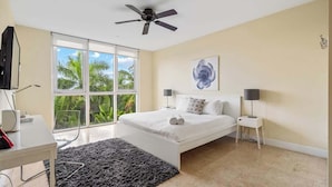 A bedroom with a bed, a ceiling fan, a window with a view of palm trees, and a small table with a lamp. - Hemingway Condo - Sunset View & steps to the beach (Deerfield Beach)