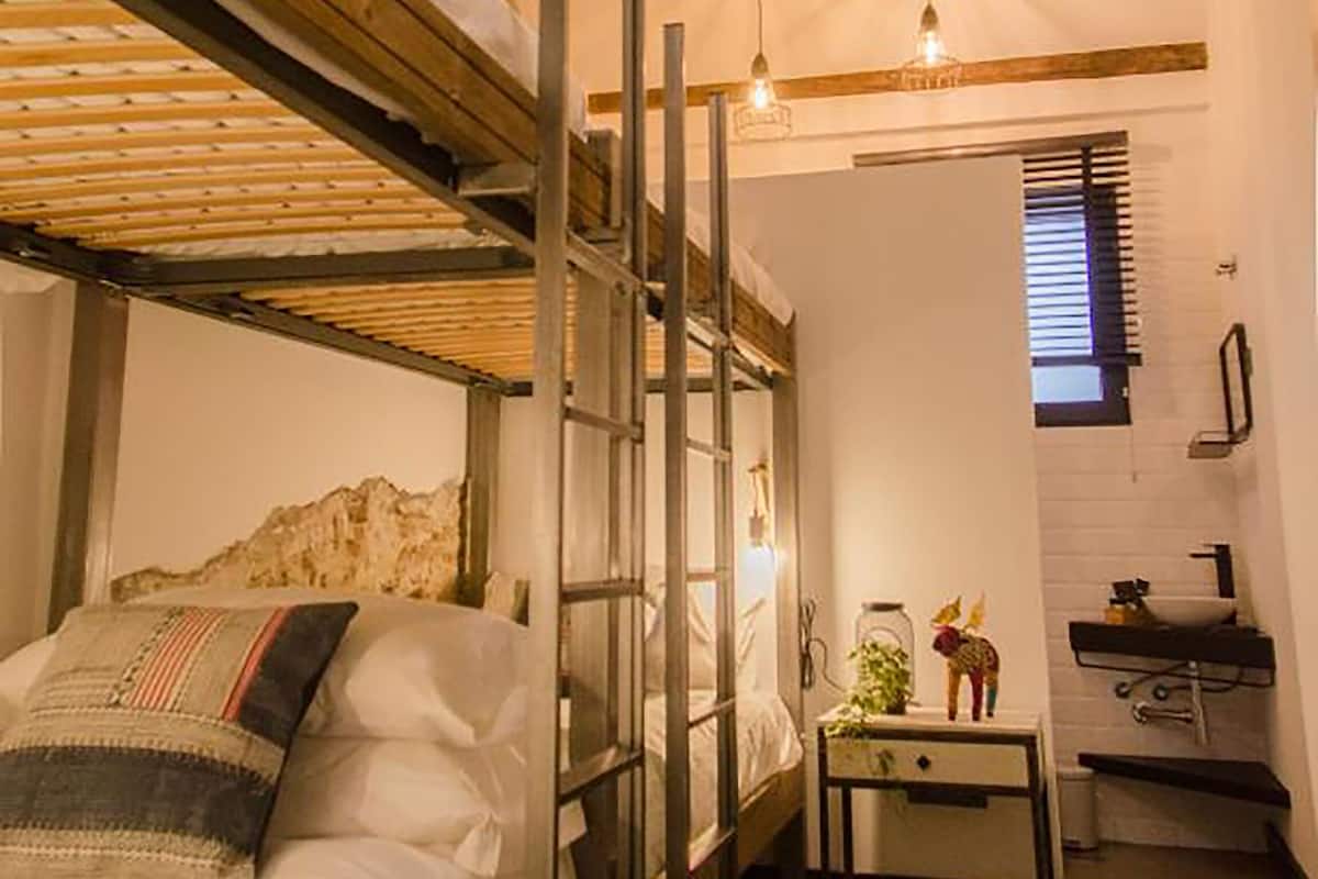 Quarto compacto com beliche, uma pequena mesa com planta e janela com persianas.