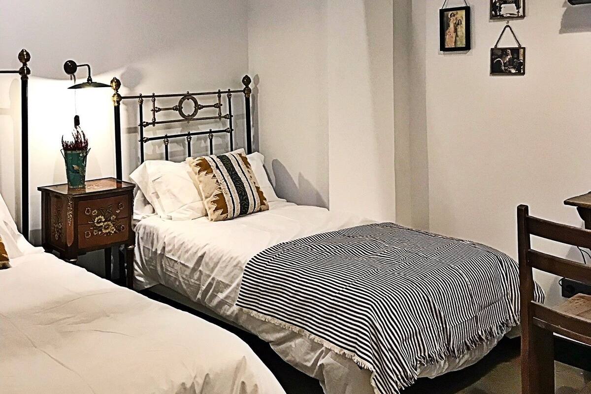 Quarto com duas camas de solteiro, cabeceira de madeira, mesa de cabeceira com abajur e uma cadeira pequena.