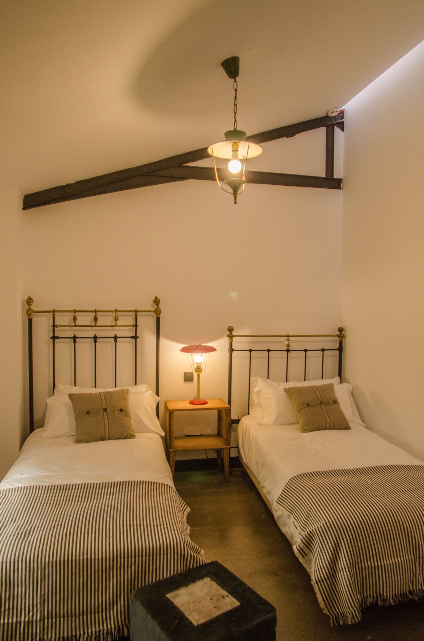 A room with two beds, each with a striped bedspread, a small table with a lamp, and a hanging light fixture.