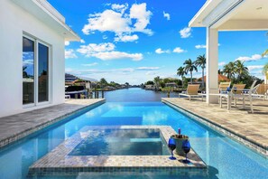 Una piscina con agua azul cristalina, rodeada de sillas de descanso y palmeras, con vistas al cielo y edificios a lo lejos.