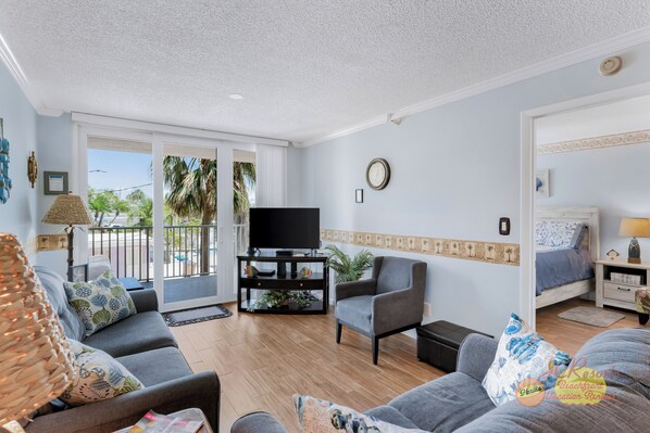 Una sala de estar con sofá, sillón, televisión y una cama en una habitación con paredes azul claro y un balcón.