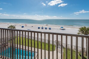 Blick auf den Strand vom Balkon mit Pool, Strandliegen und blauem Himmel.