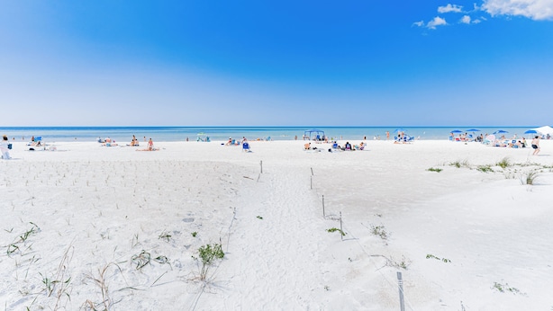 Una playa con gente tomando el sol y nadando, bajo un cielo azul despejado.