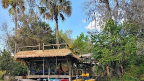 Une construction à toit de chaume dotée d’une terrasse en bois et d’un espace salon, entourée d’eau et de verdure.