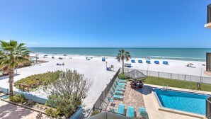 A beach view from a balcony with a swimming pool, lounge chairs, and palm trees.