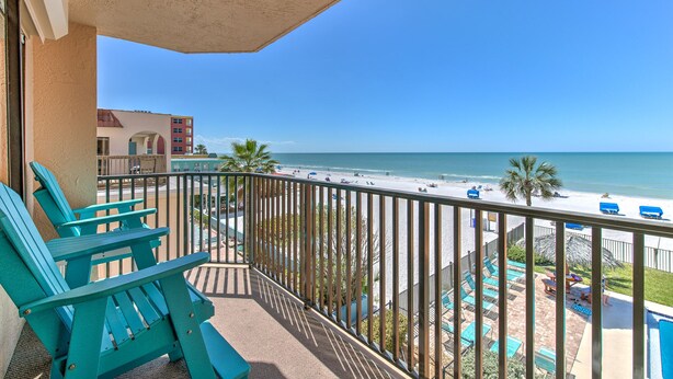A balcony with teal Adirondack chairs, a pool area with lounge chairs, and a view of the beach and ocean.