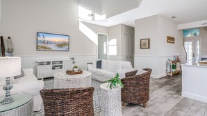 A spacious living room with a white sofa, wicker chairs, a round coffee table, and a painting of a beach scene. - Pearl Cottage Private Heated Pool Home! Newly renovated book today! (Palm Coast)