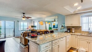 A spacious kitchen with white cabinets, a marble countertop, and a large island. There is a ceiling fan, pendant lights, and a view of the outside through large windows.