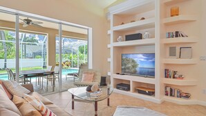 A living room with a glass coffee table, a sofa, a TV on a white shelf, and a pool view.