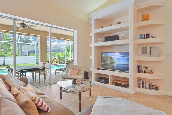A living room with a glass coffee table, a sofa, a TV on a white shelf, and a pool view.