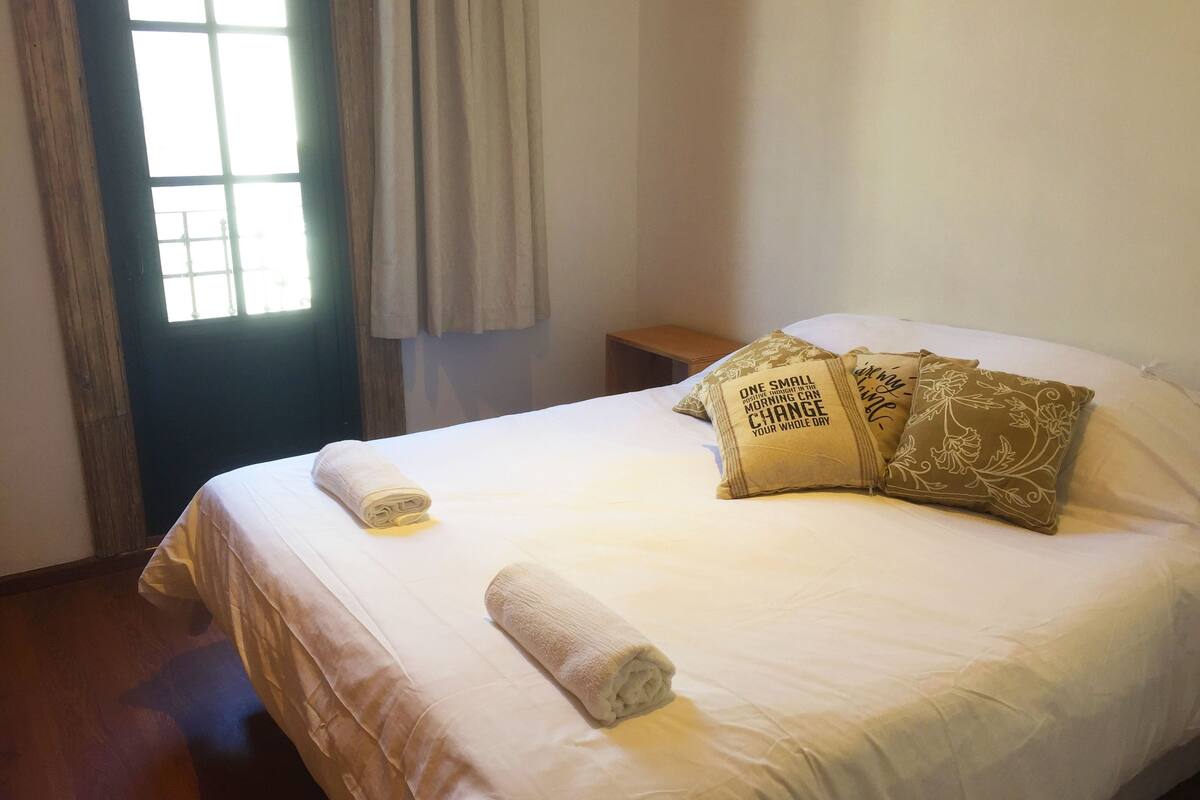 A neatly made bed with a rolled white towel, two decorative pillows with floral patterns, and a wooden nightstand beside the bed.