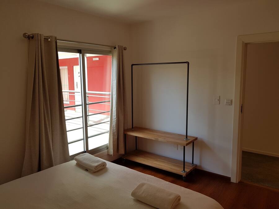 Una habitación de hotel con una cama, un estante de madera, una ventana con cortinas y vista a un edificio rojo.