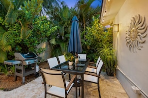 An outdoor patio with a glass table and chairs, a grill, and lush greenery. - Experience Casa del Sol: A Midcentury Tropical Getaway with Classic South Florida Vibes! (Wilton Manors)