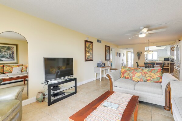 A living room with a television, sofa, coffee table, and kitchen in the background. - Oceanfront Cocoa Beach Condo w/ View: Walk to Pier (Cocoa Beach)